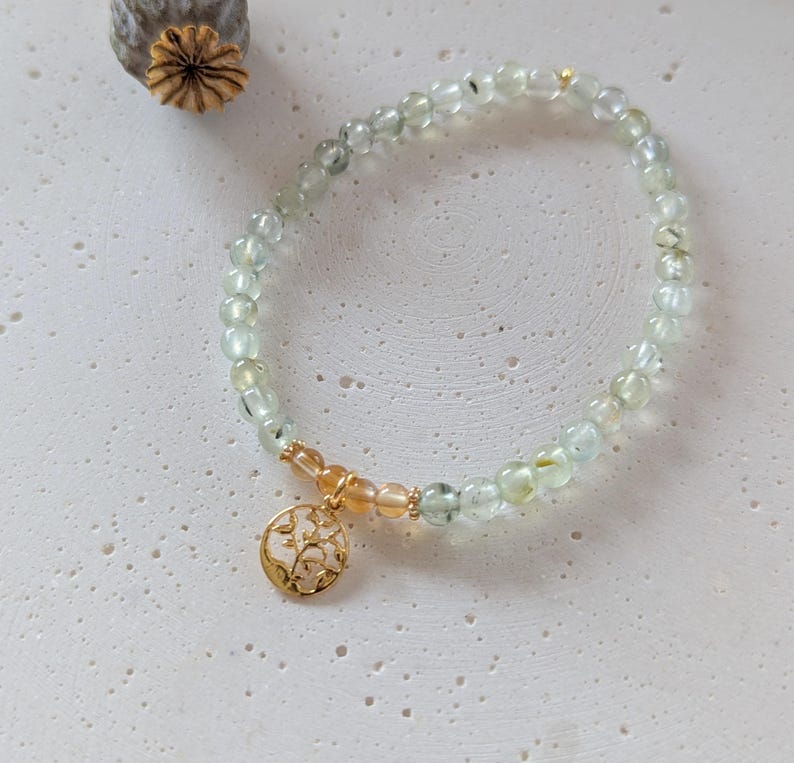 Op de afbeelding: Een delicate armband met lichtgroene en heldere kralen. Een goudkleurige bedel met een bloemmotief hangt aan de armband. De armband wordt gepresenteerd op een getextureerd, gebroken witte ondergrond. Een gedroogde zaaddoos staat op de achtergrond.
