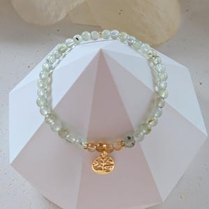 Op de afbeelding: Een delicate armband met kleine, ronde, lichtgroene kralen met gouden accenten. Een gouden bedel met een boomontwerp hangt in het midden. De armband wordt gepresenteerd op een wit geometrisch oppervlak.