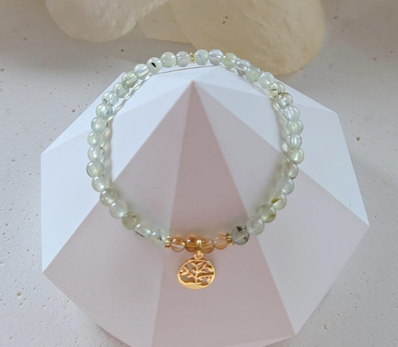 Op de afbeelding: Een delicate armband met kleine, ronde, lichtgroene kralen met gouden accenten. Een gouden bedel met een boomontwerp hangt in het midden. De armband wordt gepresenteerd op een wit geometrisch oppervlak.