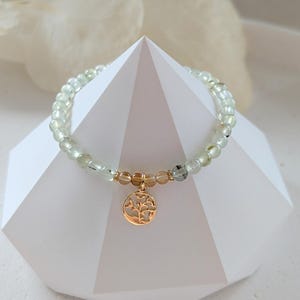Op de afbeelding: Een armband met lichtgroene en heldere kralen, geaccentueerd met goudkleurige kralen en een ronde bedel met een boomontwerp. De armband wordt gepresenteerd op een witte geometrische vorm.