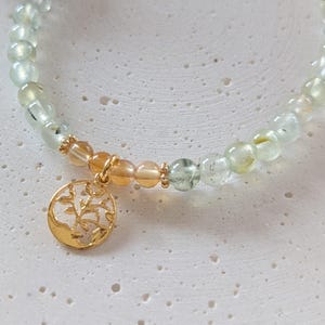 Op de afbeelding: Een armband met lichtgroene en amberkleurige edelstenen. Een goudkleurige bedel met een boomontwerp hangt eraan. De armband wordt getoond op een getextureerd, gebroken witte ondergrond.