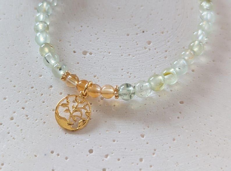 Op de afbeelding: Een armband met lichtgroene en amberkleurige edelstenen. Een goudkleurige bedel met een boomontwerp hangt eraan. De armband wordt getoond op een getextureerd, gebroken witte ondergrond.