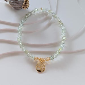 Op de afbeelding: Een delicate armband met lichtgroene en heldere kralen, geaccentueerd met goudkleurige afstandhouders en een gouden hanger. De hanger heeft een boomontwerp. De armband is een stijlvol accessoire.
