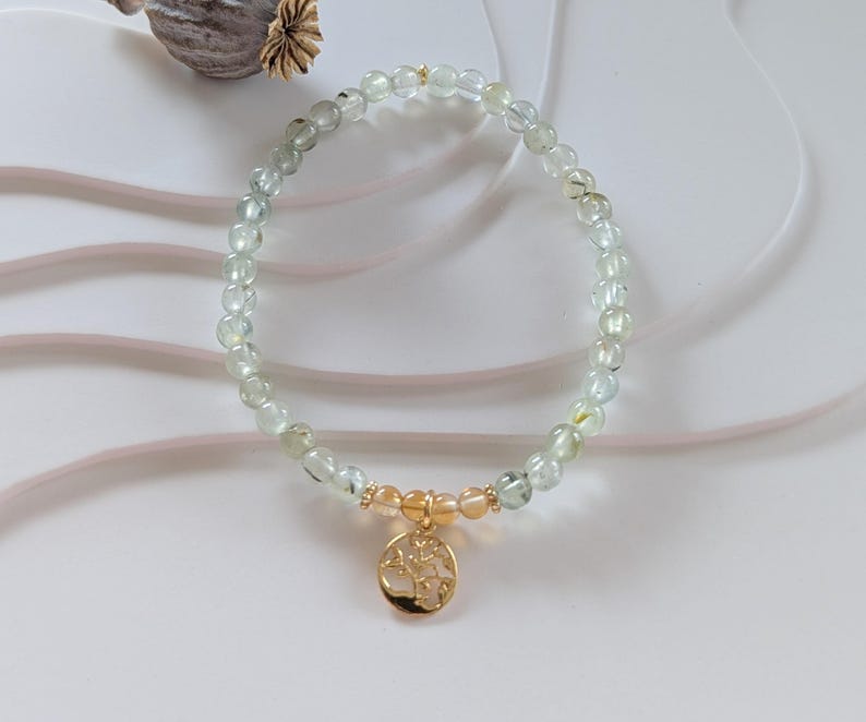 Op de afbeelding: Een delicate armband met lichtgroene en heldere kralen, geaccentueerd met goudkleurige afstandhouders en een gouden hanger. De hanger heeft een boomontwerp. De armband is een stijlvol accessoire.