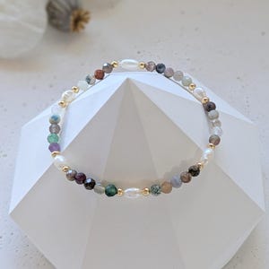 Könnte beinhalten: Ein mehrfarbiges Perlenarmband mit goldenen Akzenten. Das Armband besteht aus verschiedenen Steinen, darunter weiß, grau, grün und lila.