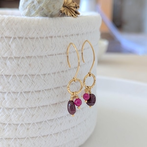 Könnte beinhalten: Ein Paar goldfarbene Ohrringe mit Haken-Design. Jeder Ohrring hat einen kleinen gehämmerten Ring mit einer Ansammlung von drei Edelsteinen: zwei runde, leuchtend rosa Steine und ein ovaler, dunkelroter Stein.