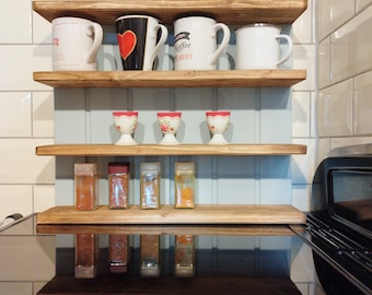 May include: Three wooden shelves with a light blue background. The top shelf has four mugs, one with a red heart design, one with the word "Kaffee" on it, and two white mugs. The middle shelf has four white egg cups with red and white floral designs. The bottom shelf has six glass spice jars with various spices.