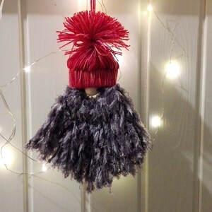 Puede incluir: Un adorno de pompon de hilo gris con un gorro de punto rojo y un pompon en la parte superior. El adorno está colgado de una cadena de luces blancas de hadas sobre un fondo de madera blanca.