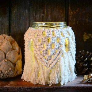 Pode incluir: Um abajur de macramé branco com franjas, cobrindo um frasco de vidro com uma luz quente dentro. O abajur está sobre uma superfície de madeira com uma pinha e uma pedra decorativa.