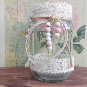 Puede incluir: Un tarro de vidrio transparente decorado con encaje blanco y cuentas rosas y blancas. El tarro está atado con una cinta rosa y un cordel. El tarro está sentado sobre una superficie de madera.