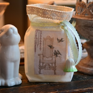 Könnte beinhalten: Eine weiße Keramik-Hasenfigur mit einer Stofftasche mit Spitzenbesatz, die mit einem Blumendesign und Vögeln verziert ist. Die Tasche hat einen grün-weißen Glöckchenanhänger.