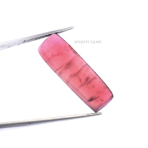 May include: A rectangular, translucent pink gemstone with visible internal inclusions and a smooth surface, held by silver-toned metal tweezers. The text HIT&FIT GEMS is printed in small grey letters near the top right of the pink stone.
