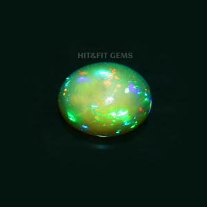 May include: An oval-shaped cabochon opal gemstone featuring a translucent body with vibrant flashes of green, blue, and orange play-of-color. The text HIT&FIT GEMS is printed in light gray above the stone against a solid black background.