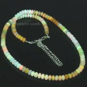 May include: A necklace composed of small, colorful opal beads. The beads transition in color from white to yellow, orange, and green. The necklace is strung on a silver chain with a lobster clasp. The text "HIT&FIT GEMS" is visible.