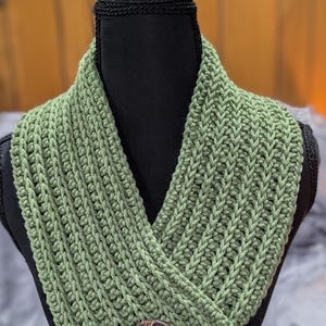 May include: A sage green crochet cowl scarf with a textured stitch pattern. The scarf is secured with a round, brown coconut shell button. The cowl is displayed on a black mannequin head against a neutral background.