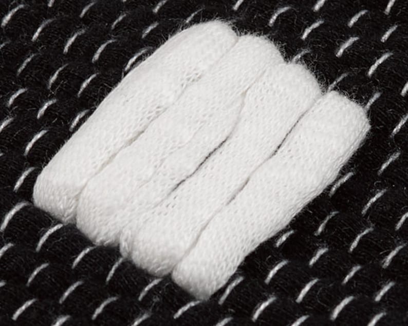May include: Close-up of a white fabric patch sewn onto a black fabric background. The patch is made of several layers of fabric, creating a textured look.
