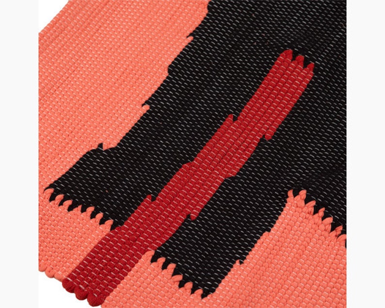 May include: A woven rug with a geometric pattern in shades of coral, red, and black. The rug has a textured surface and a fringed edge.