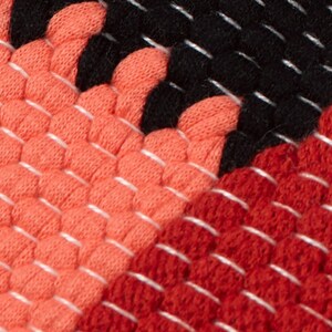 May include: Close-up of a woven fabric with a geometric pattern in shades of red, orange, and black. The fabric has a textured, bumpy surface.