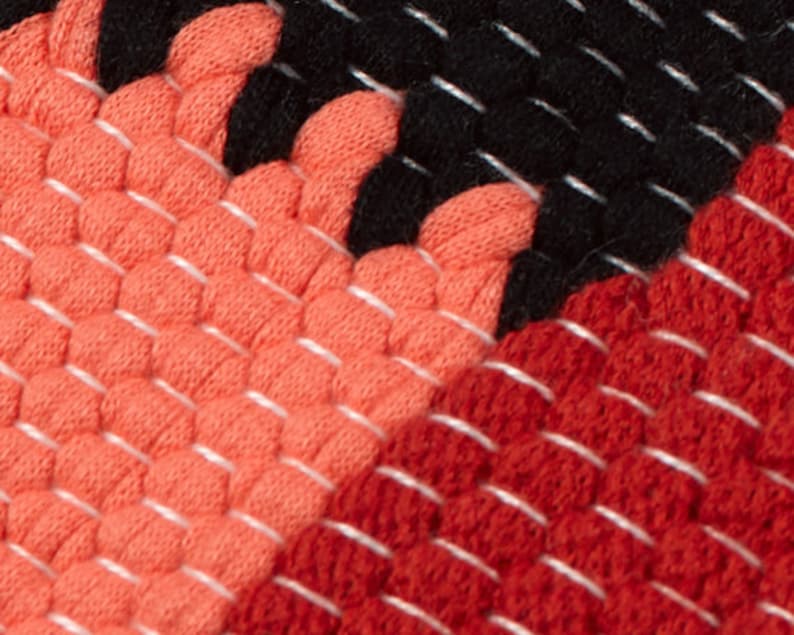 May include: Close-up of a woven fabric with a geometric pattern in shades of red, orange, and black. The fabric has a textured, bumpy surface.
