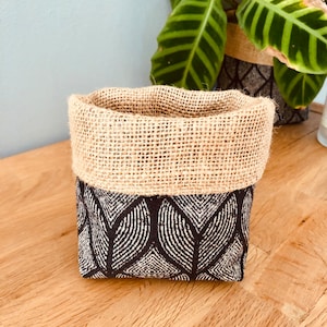 Può includere: Un piccolo cesto di stoccaggio di forma quadrata con una parte superiore in juta e un motivo geometrico bianco e nero sul fondo. Il cesto è appoggiato su una superficie di legno.