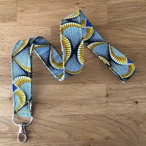 Puede incluir: Un cordón con estampado y cierre plateado. La tela presenta un diseño repetitivo de formas de abanico amarillas, azules y negras. El cordón está sobre una superficie de madera.