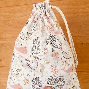 May include: A white drawstring bag with a mermaid and sea life pattern. The bag is made of cotton fabric and has a white drawstring.