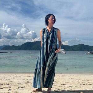 May include: A sleeveless, floor-length dress with vertical stripes in shades of blue and silver. The dress has a V-neck and a tiered skirt, worn on a sandy beach with a backdrop of ocean and sky.