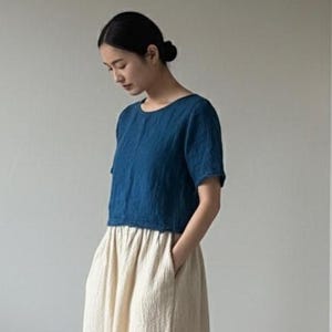 May include: A woman wearing a two-piece outfit. The top is a short-sleeved, dark blue shirt. The bottom is a long, cream-colored skirt with pockets. She is wearing sandals.