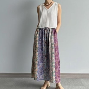 Puede incluir: Una mujer con una camiseta sin mangas blanca y una falda larga estampada. La falda presenta diseños florales morados, azules y rosas. También lleva un collar y sandalias.