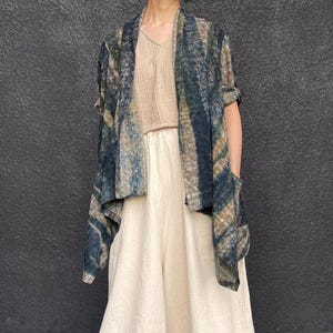 May include: A draped, open-front cardigan in shades of blue, green, and brown, with a textured pattern. The cardigan is paired with a beige top and wide-leg, cream-colored pants. The outfit has a relaxed, bohemian style.