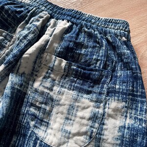May include: Close-up of blue and white patterned shorts. The shorts have an elastic waistband and a single pocket. The fabric has a distressed, tie-dye appearance. The color palette includes shades of blue and white.
