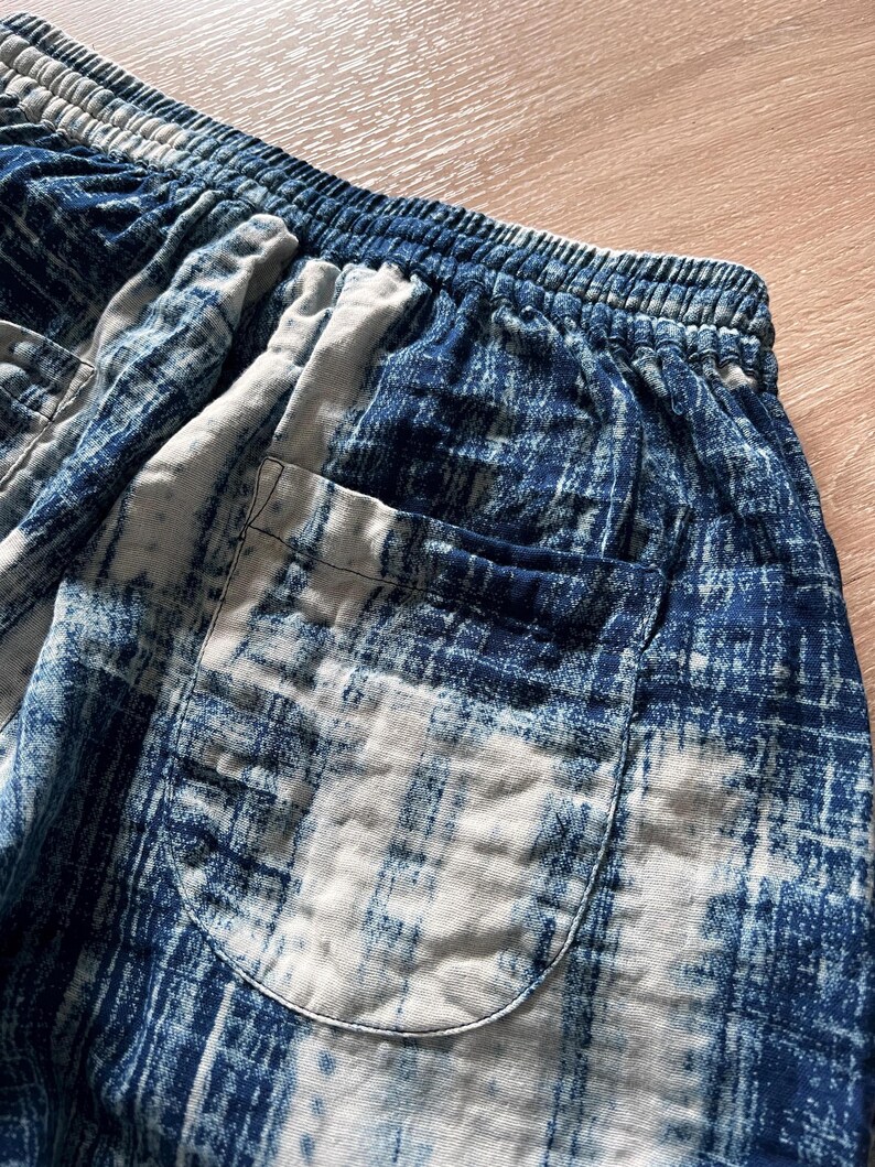 May include: Close-up of blue and white patterned shorts. The shorts have an elastic waistband and a single pocket. The fabric has a distressed, tie-dye appearance. The color palette includes shades of blue and white.