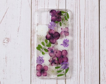 May include: A clear phone case with a pressed flower design. The flowers are purple, white, and green.
