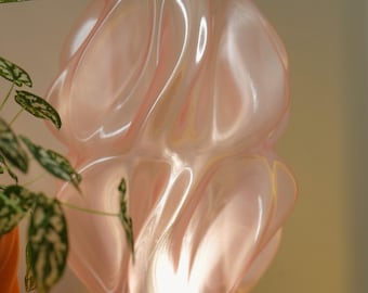 May include: A soft pink, sculptural table lamp with a wavy, organic design. The lamp sits on a red base and emits a warm glow. The lamp is approximately 10 inches tall. A houseplant is visible in the background.