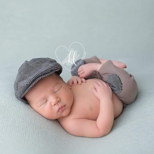 以下が含まれることがあります： グレーのチェック柄の帽子と、チェック柄のパッチが付いた茶色のズボンを履いた、眠っている新生児。赤ちゃんは水色の毛布の上に横たわっています。画像は柔らかくパステル調です。