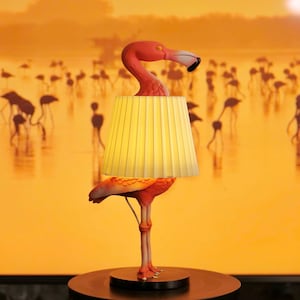 Może przedstawiać: Lampa stołowa w kształcie flaminga z żółtym abażurem. Lampa jest pomarańczowa i różowa, z okrągłą czarną podstawą. Tło przedstawia scenę zachodu słońca z sylwetkami flamingów.