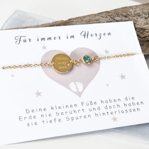 Könnte beinhalten: Ein goldenes Kettenarmband mit einem kleinen runden Anhänger, auf dem "Always in my heart" steht, und einem hellblauen Kristall. Das Armband liegt auf einer weißen Karte mit dem Text "Für immer im Herzen" und einem Gedicht über kleine Füße, die eine große Wirkung hinterlassen.