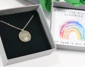 Könnte beinhalten: Eine silberne Halskette mit einem runden Anhänger, auf dem "Vince" steht und ein kleiner Schulbus eingraviert ist. Die Halskette befindet sich in einer grauen Geschenkbox mit einer Karte, auf der "Hurra, ich bin Schulkind" steht und ein Regenbogen abgebildet ist.