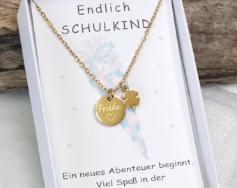 Könnte beinhalten: Eine goldfarbene Halskette mit einem runden Anhänger, der mit dem Namen "Frieda" und einem Herz graviert ist, sowie einem Kleeblatt-Charm. Die Halskette wird in einer weißen Schachtel mit deutschem Text präsentiert: "Endlich SCHULKIND" und "Ein neues Abenteuer beginnt. Viel Spaß in der Schule!"