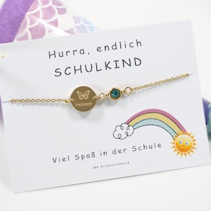 Könnte beinhalten: Goldarmband mit einem runden Anhänger, der mit dem Namen "Hannah" und einem Schmetterlingsmotiv graviert ist. Der Anhänger ist an einer zarten goldenen Kette mit einem kleinen blauen Edelstein befestigt. Das Armband wird auf einer weißen Karte mit dem Text "Hurra, endlich SCHULKIND" und "Viel Spaß in der Schule" präsentiert.