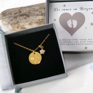 Könnte beinhalten: Eine goldene Halskette mit einem Sternanhänger und einem runden Anhänger, der mit einer Konstellation und Fußabdrücken graviert ist. Die Halskette befindet sich in einer grauen Geschenkbox mit einer weißen Karte, auf der "Für immer im Herzen" steht, und einem Zitat über Fußabdrücke.