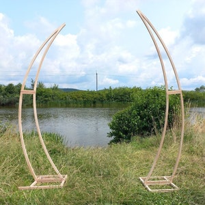 Könnte beinhalten: Zwei Holzbögen aus Naturholz, die auf einer grasbewachsenen Wiese in der Nähe eines Teichs stehen. Die Bögen sind gebogen und haben ein schlichtes Design.