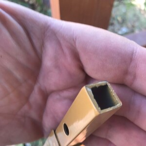 May include: Close-up of a gold metal rod with a square hollow center. The rod has a small hole drilled through it.