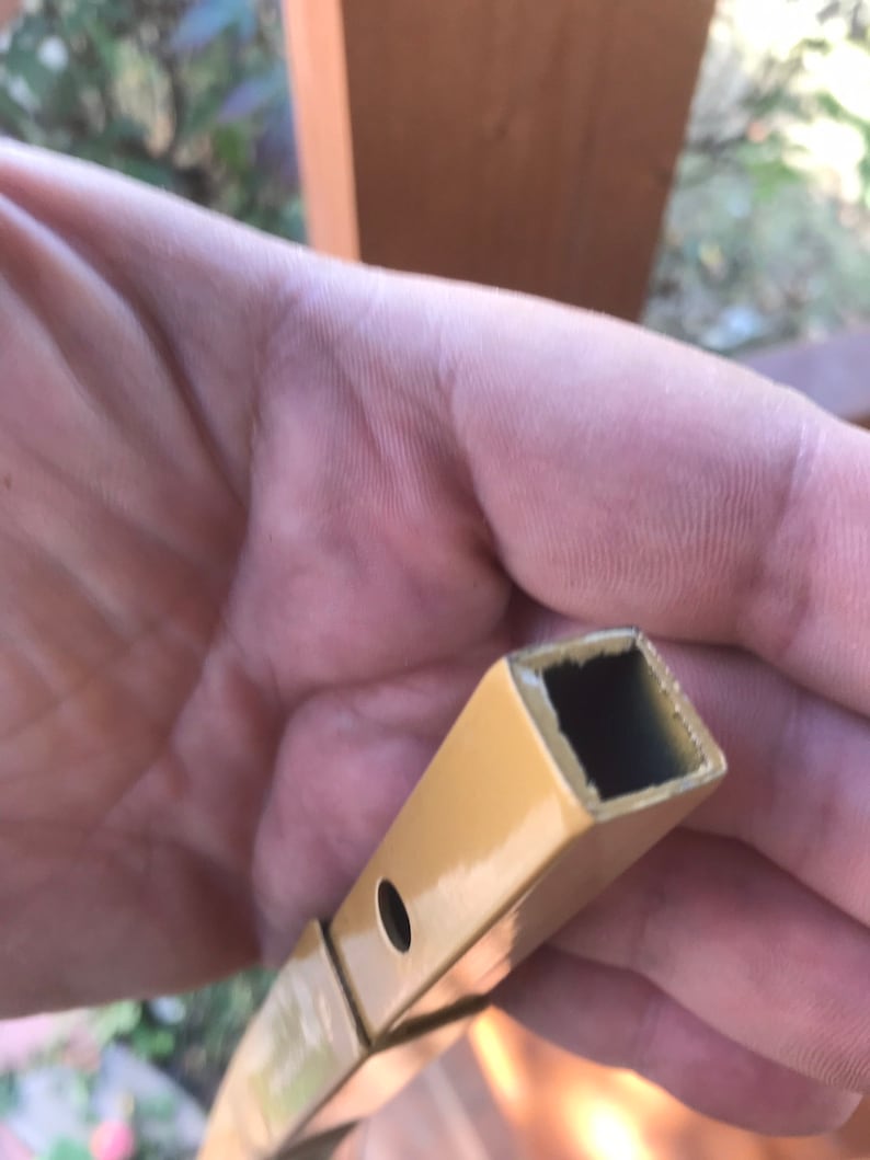May include: Close-up of a gold metal rod with a square hollow center. The rod has a small hole drilled through it.