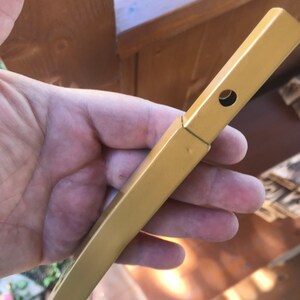May include: A gold metal rod with a hole drilled through it. The rod is held in a hand.