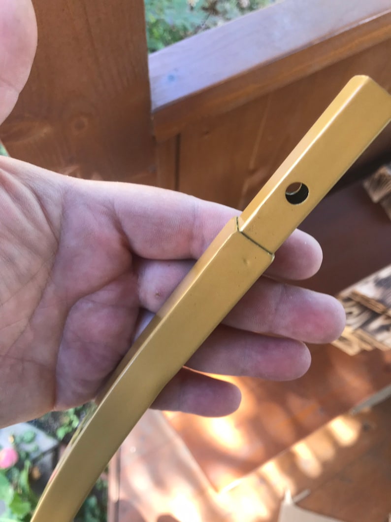 May include: A gold metal rod with a hole drilled through it. The rod is held in a hand.