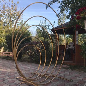 May include: A gold metal archway made of five concentric circles. The archway is standing on a brick patio in front of a wooden gazebo.