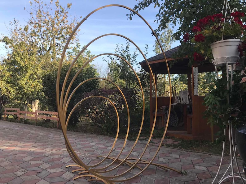 May include: A gold metal archway made of five concentric circles. The archway is standing on a brick patio in front of a wooden gazebo.