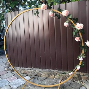 May include: A gold metal hoop arch decorated with a garland of pink artificial roses and green leaves. The arch is freestanding and has a simple, modern design.