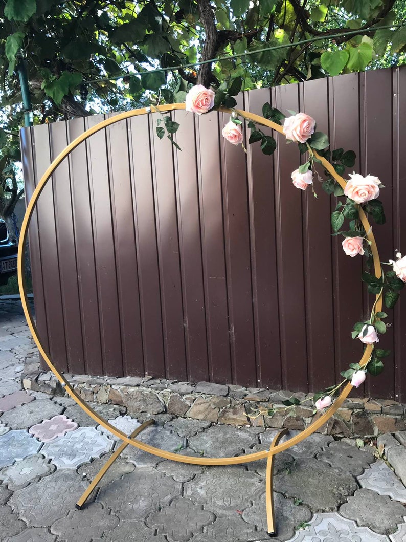 May include: A gold metal hoop arch decorated with a garland of pink artificial roses and green leaves. The arch is freestanding and has a simple, modern design.